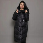 Parka matelassée femme conception longue et épaisse avec capuche, de couleur noire, portée par une femme. Sur un fond marron et gris.