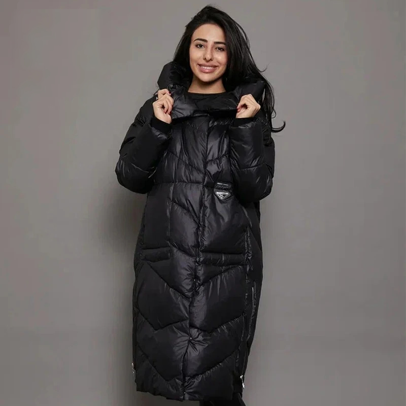 0-a85bc8.jpeg Parka matelassée femme conception longue et épaisse avec capuche, de couleur noire, portée par une femme. Sur un fond marron et gris.