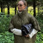 Parka matelassée femme avec col haut et manches courtes de couleur kaki. Porté par une femme avec lunettes de soleil et un sac dans la main. Sur un fond style forêt verdoyante.